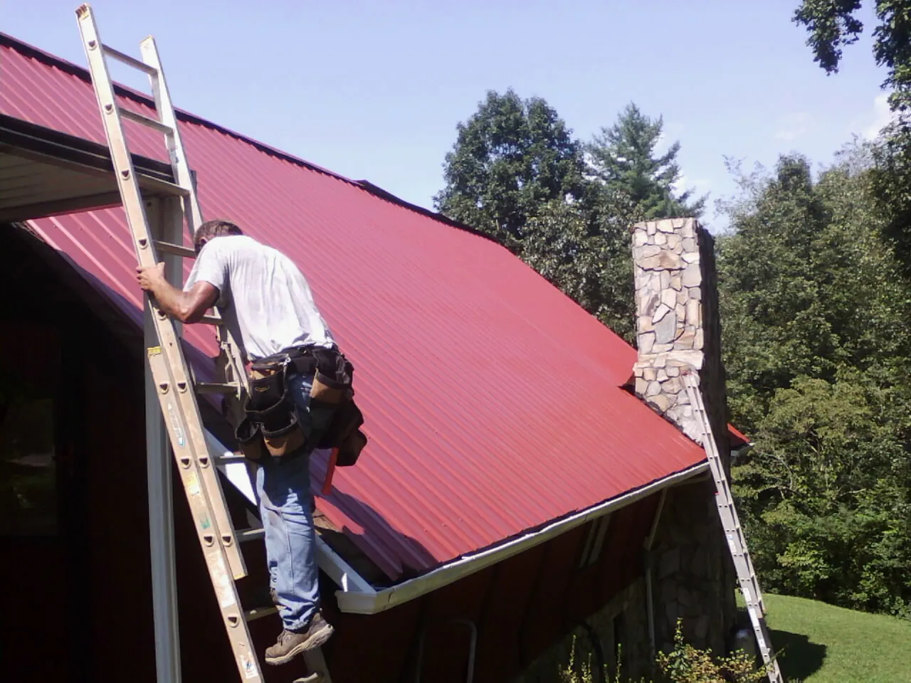 Roofer on a ladder beside a red metal roof and stone chimney — Soffit & Fascia Repair by Roofing Brothers in Clifton Heights